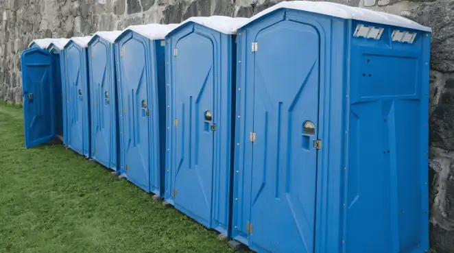 Row of blue portable toilets lined up on grass against a stone wall for event sanitation in Jersey City, NJ
